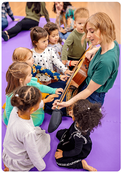 A group of children gathered around their Music Together teacher, who is playing a guitar